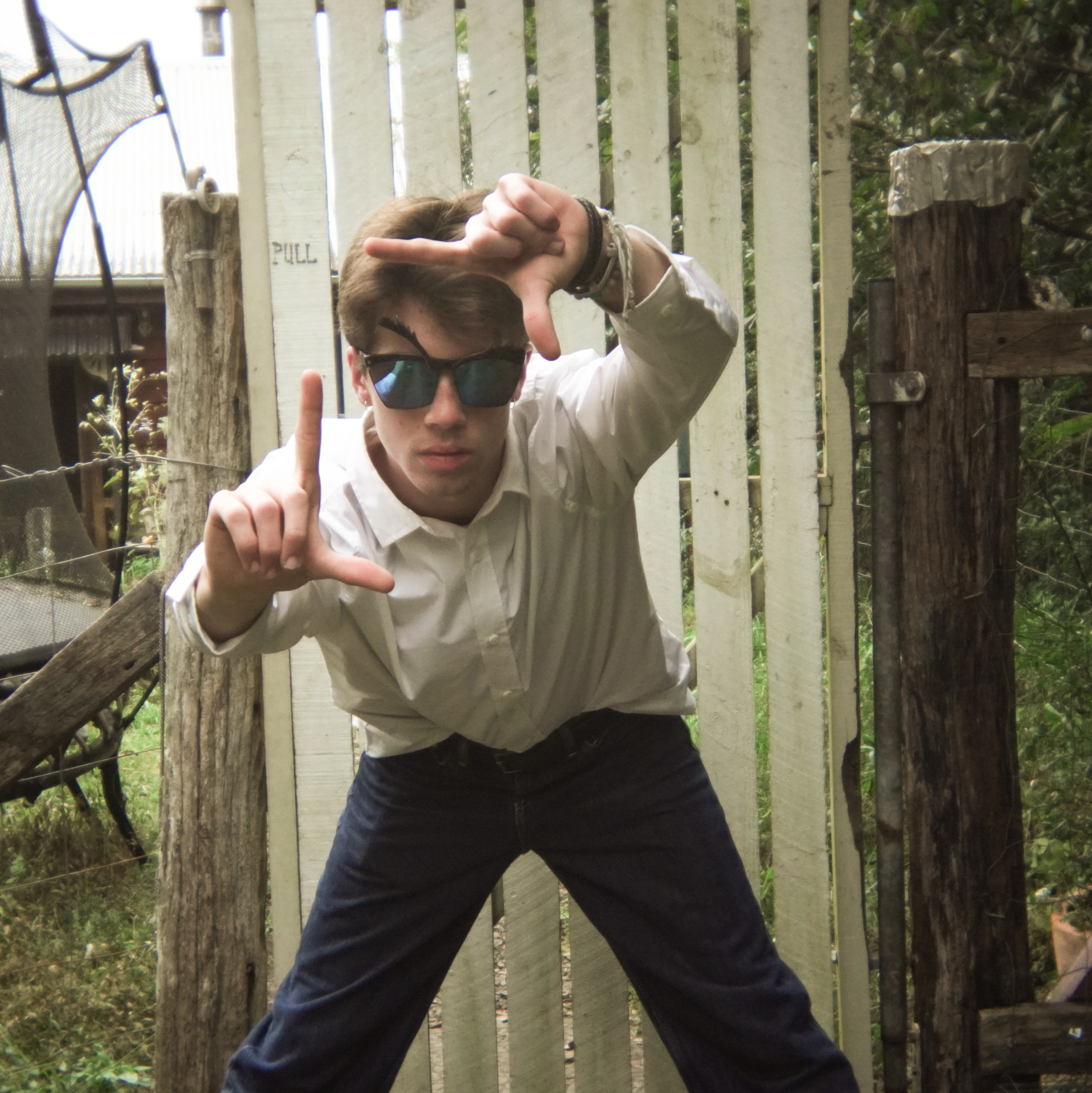 The image shows Luke Dunlop outdoors, wearing a white shirt and reflective sunglasses, striking an iconic Willem Dafoe pose with his fingers framing a scene.